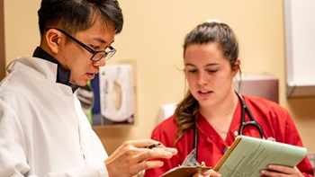 Two health sciences students wear lab coats and look at a clipboard.
