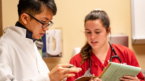 Two health sciences students wear lab coats and look at a clipboard.