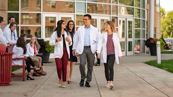 Students walking and talking outside the medical school. They are all wearing professional attire and white medical coats.