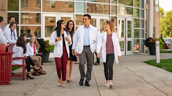 Students walking and talking outside the medical school. They are all wearing professional attire and white medical coats. 