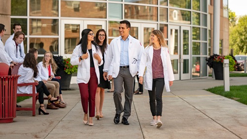 Students walking and talking outside the medical school. They are all wearing professional attire and white medical coats.