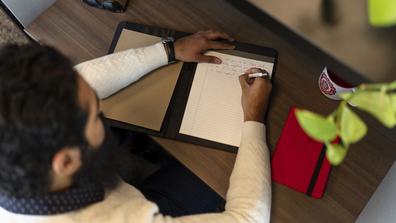 A student writes in their notebook with a USD padfolio next to them. A student writes in their notebook with a USD padfolio next to them.