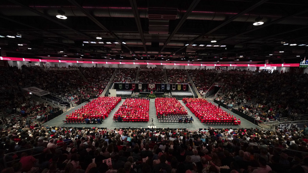 A photo of the University of South Dakota undergraduate commencement ceremony.  A photo of the University of South Dakota undergraduate commencement ceremony. Thousands of students wearing red gowns sit as they listen to the speaker.