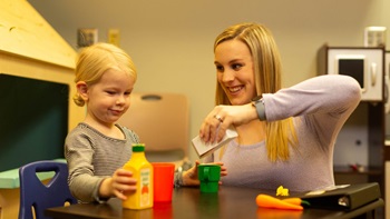 A woman plays with toy food with a child at a table.