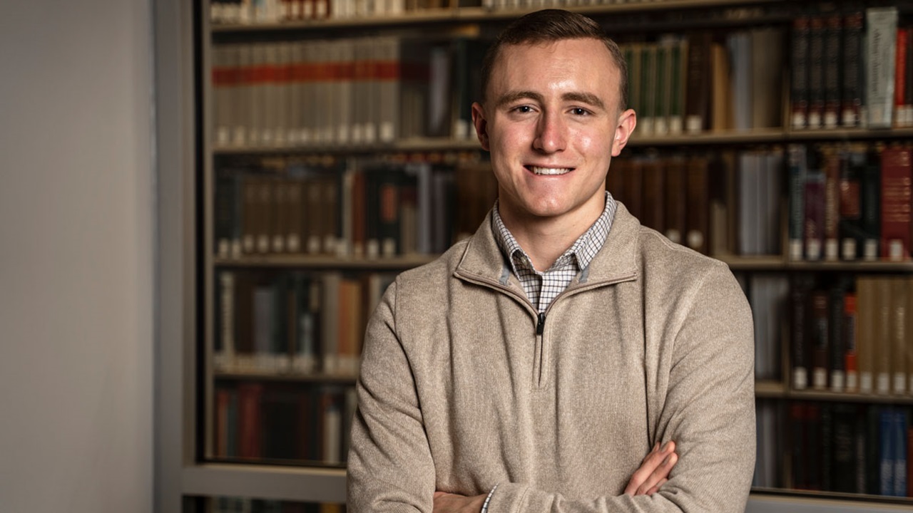 Caleb Swanson standing in a library with his arms crossed and looking at the camera. Caleb Swanson standing in a library with his arms crossed and looking at the camera.