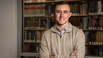 Caleb Swanson standing in a library with his arms crossed and looking at the camera.