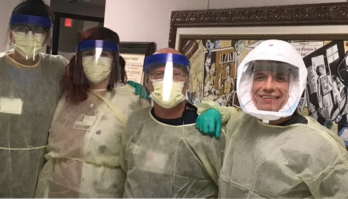 A group of people wear full body personal protective equipment and pose with their arms around each other.