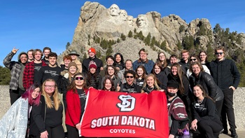 The USD Chamber Singers at Mount Rushmore. They are together as a group and the students in front hold the USD flag.
