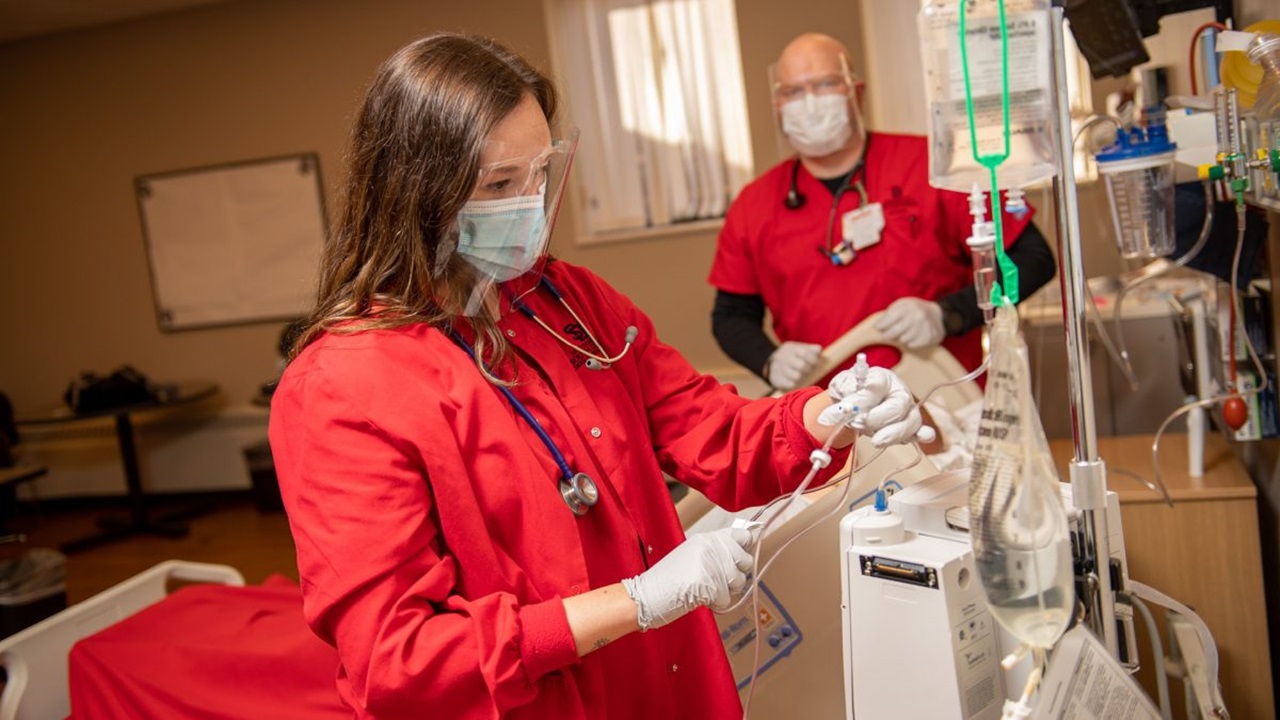 Nursing student in a red coat looks at an IV in a hospital room.
