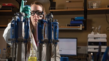 A USD student fills a beaker in a lab wearing a white coat and goggles.