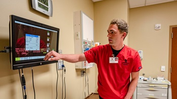 A USD nursing student in red scrubs points at a medical display monitor in a hospital room.