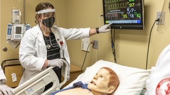 A USD student stands over a manikin, reading its chart and explaining something to their team.