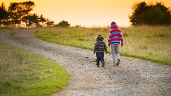 Two children walk away on a gravel road. There is grass on both sides of the road, and the sun is setting.