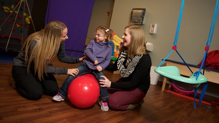 Two women help a child on an exercise ball.