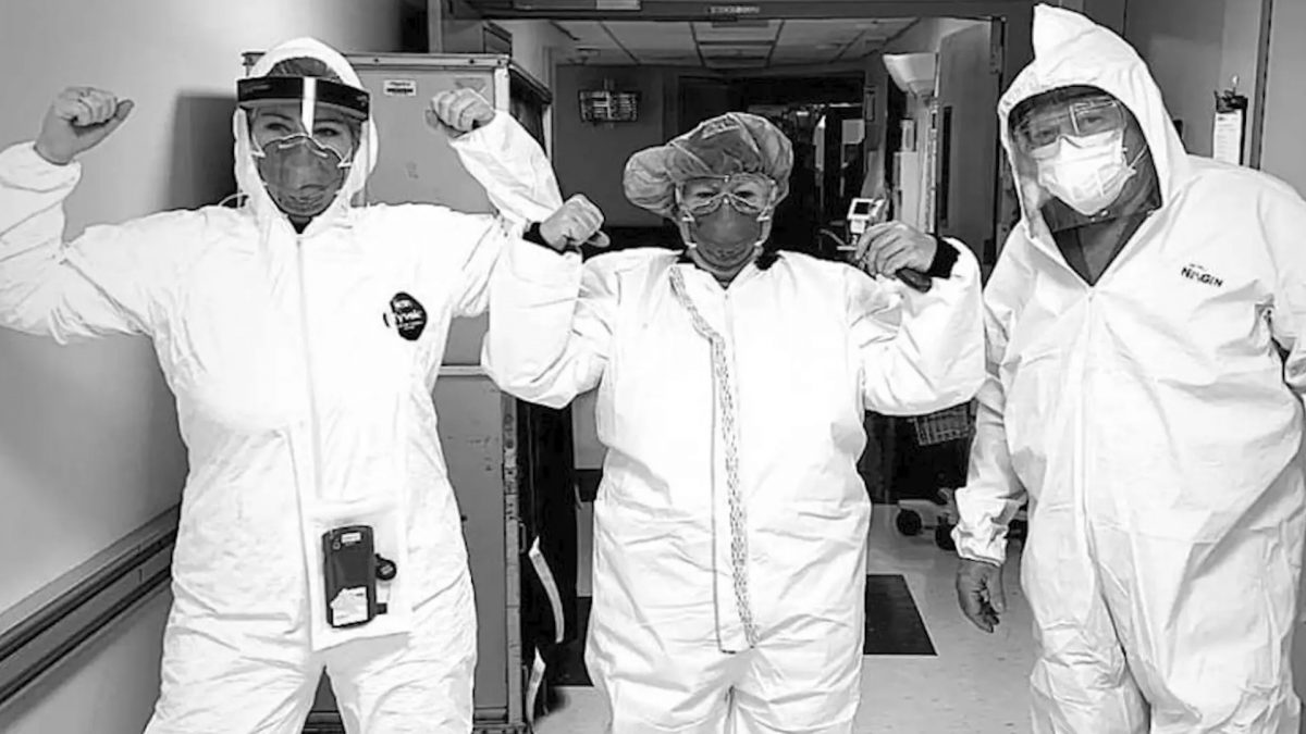 Three people in hazmat suits and masks flex their arm muscles and pose in a hospital.
