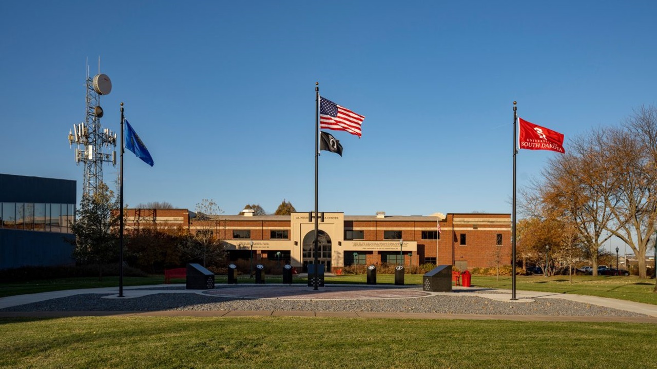 The South Dakota state flag, American flag, prisoner of war flag and University of South Dakota flag fly over the Patriot's Plaza on USD's campus. The sky is blue.