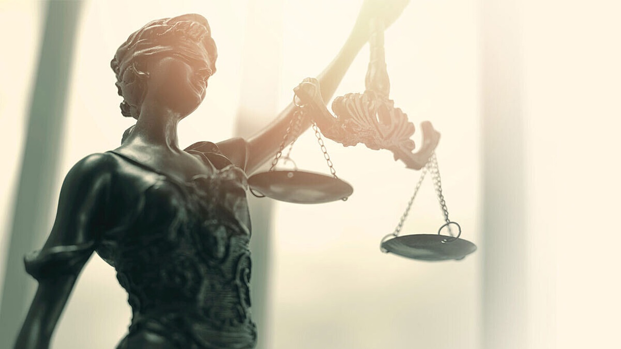 A dark gray statue holds a balancing scale. Light shines on the statue.