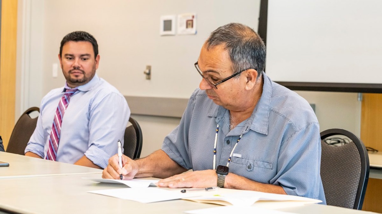 A person signs a document at a table while another person sits by and watches.