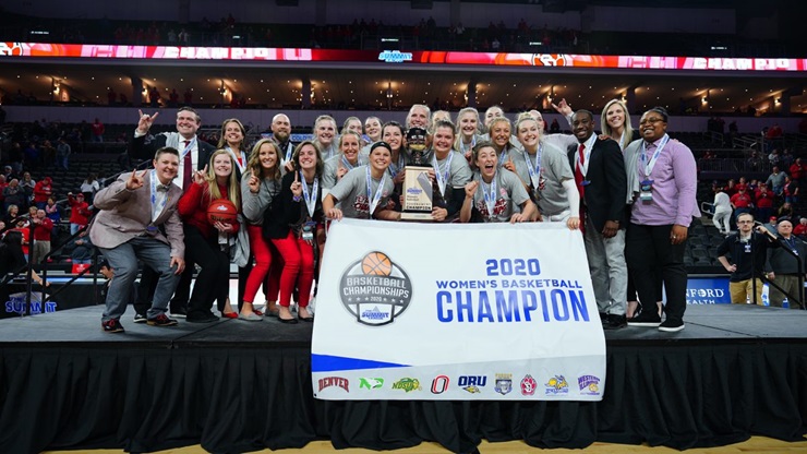 The 2020 USD women's basketball team holds up the 2020 Summit League champions poster and trophy and poses on a stage. 