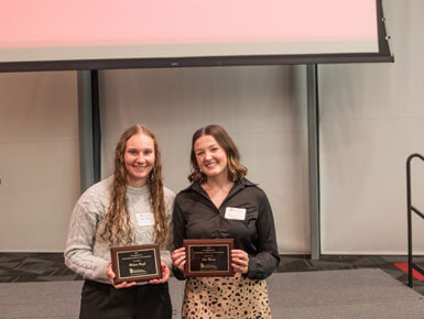 Two Beacom School of Business alumnae stand in front of the stage and screen, holding their award plaques and smiling at the camera.