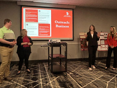 Four CBC presenters stand at the front of a room discussing an “Outreach: Business” slide during a group presentation.
