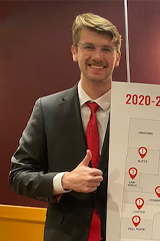 A student wears a suit and a red tie and holds up a posterboard. He has a thumbs up.
