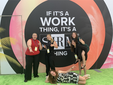 USD students take a photo in front of a black, pink, yellow, and orange sign that says, "If it's a work thing, it's a SHRM thing."