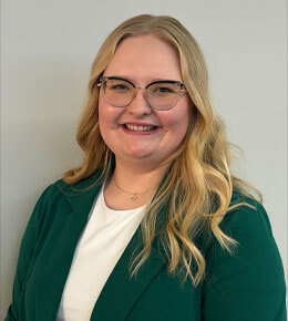 Makayla Van Voorst poses for a professional photo. She is wearing glasses, with a white shirt and green cardigan. She is smiling at the camera.
