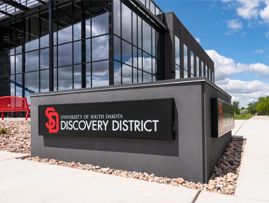 The USD Discovery District's building sign stands on the corner of the campus, with the Discovery District building in the background, with sunny skies and green grass surrounding it.