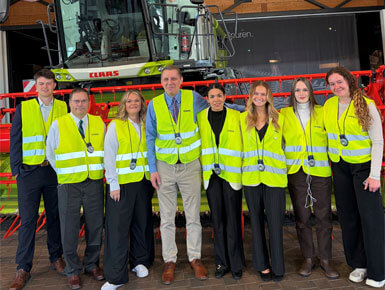 Members of the Trade Mission Trip stand in a construction zone or an industrial facility. They are wearing high-visibility yellow safety vests stand in front of a large agricultural machine.