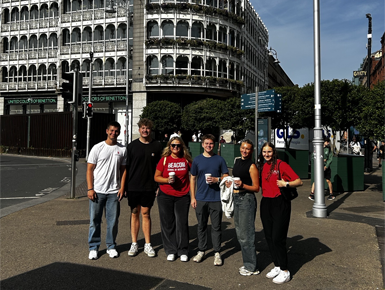 A group of six young adults standing on a city street in front of a historic building with arched windows. They are casually dressed and smiling at the camera.