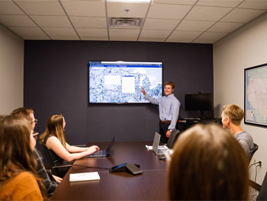 A male USD student stands at the head of a conference table presenting a digital map on a large monitor to a group of seated students.