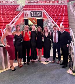 Katie Gellerman, third from the left, stands with other USD students in formal and professional attire, looking at the camera and smiling, with red bleachers in the background.