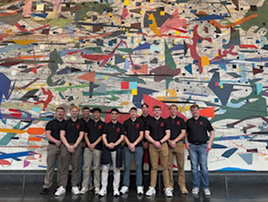 A group of twelve young men wearing matching black polo shirts and khaki or jeans posing in front of a large, colorful abstract mural.