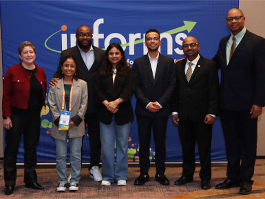 Students and mentors stand together in front of a blue backdrop that says "INFORMS" in white lettering.