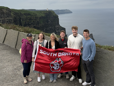 USD Beacom School of Business students and their trip leader stand on the Cliffs in Ireland, holding a red USD banner that reads, "We are South Dakota."