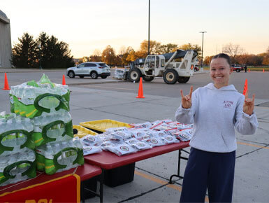 A student stands next to a table, giving the Coyote hand signal. The table is filled with bottles of water and snacks.