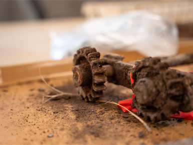 Close-up of a heavily rusted metal gear and shaft lying on a dusty wooden surface in the new USD ARCHLAB, with dirt and debris scattered around and a small piece of red plastic or tape attached nearby.