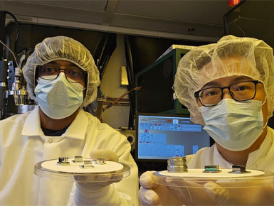 Two researchers wearing lab coats, masks, and hair nets hold circular trays with small semiconductor components in a laboratory setting.