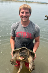 Grant Budden stands in a river and holds a large turtle in his hands. 