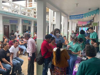 A large group of patients waits for services in a waiting room at a clinic in Guatemala. Some of the patients talk to audiology professionals in scrubs.