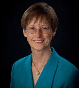 Diane Meier poses for her professional photo, which is set against a black backdrop. She is wearing a teal blazer, a necklace and glasses, and smiling at the camera.