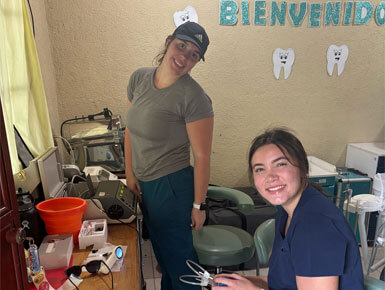 Two USD students, who are wearing scrub-like attire, are sitting and standing in a small dental office, with a sign that says, "BIENVENIDO" and tooth illustrations on the wall in the background.
