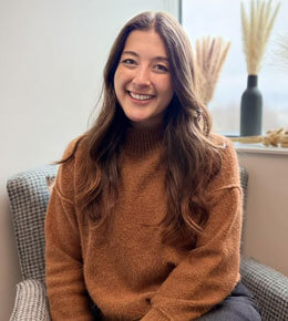 Ellen Knowles sits on a chair in the CAST Lab, posing for a picture. She is wearing a mustard-yellow sweater and smiling at the camera.