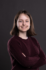 Oleksandra Lukina stands against a dark gray background and smiles for a photo with her arms crossed.