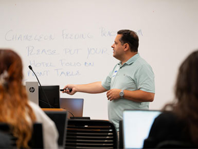 Associate Professor of Biology Christopher Anderson, Ph.D., stands in front of the STEM workshop participants, pointing toward a whiteboard.