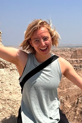 Olivia Roberts smiles wide and lifts her arms to the sides with excitement. She is outside with a clay and sandy canyon background.