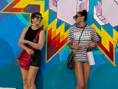 Mural On The Wall artists Amber Hansen and Reyna Hernandez stand in front of their mural in downtown Vermillion. They are looking at each other and smile. 