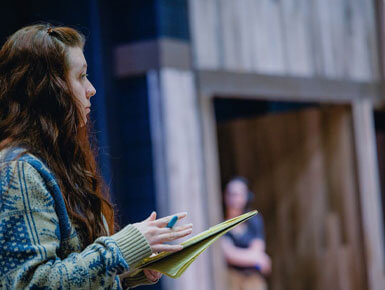 A focused director from the USD Department of Theatre, with long dark hair, gestures while holding a notebook, speaking onstage near a wooden doorway; a blurred person stands behind her.
