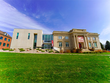 A wide-angle view shows the exterior of the National Music Museum, which is a grand, classical-style stone museum building with a modern glass entrance under a bright blue sky.